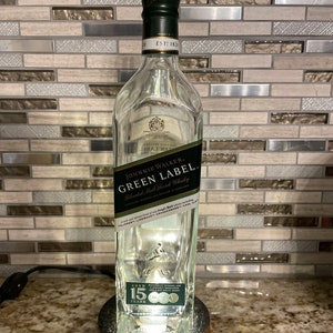 May include: A clear glass bottle with a green label that reads "JOHNNIE WALKER. GREEN LABEL. Blended Malt Scotch Whisky". The bottle is lit from the bottom and has a wooden base. The label also includes the text "AGED 15 YEARS".