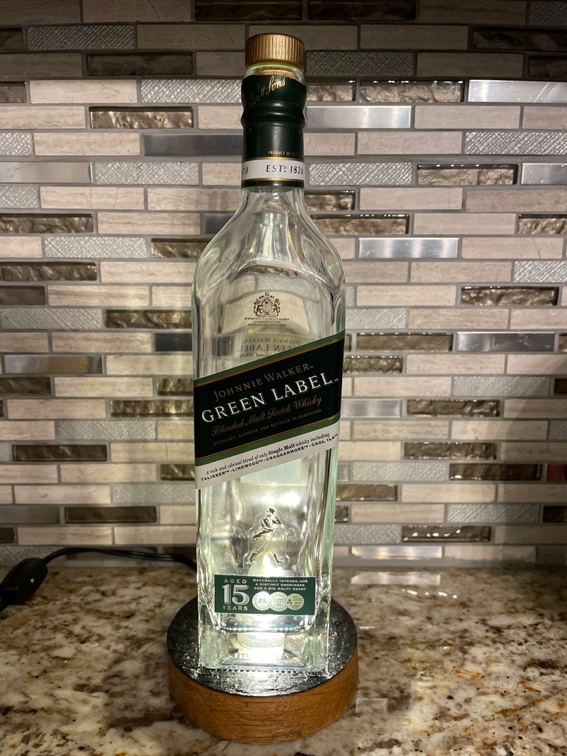 May include: A clear glass bottle with a green label that reads "JOHNNIE WALKER. GREEN LABEL. Blended Malt Scotch Whisky". The bottle is lit from the bottom and has a wooden base. The label also includes the text "AGED 15 YEARS".