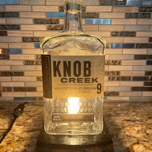 Może przedstawiać: Lampa wykonana z przetworzonej butelki po bourbonie Knob Creek. Przezroczysta szklana butelka ma etykietę z nazwą marki i szczegółami produktu. Lampa zawiera czarną podstawę i żarówkę w środku.