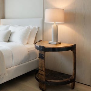 May include: A bedside table made from a repurposed wooden barrel, featuring a round top and a lower shelf. A white lamp with a beige lampshade sits on the table. The room has a bed with white linens and light-colored wood flooring.