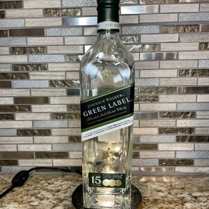 May include: A clear glass bottle with a green label that reads "Johnnie Walker Green Label Blended Malt Scotch Whisky" and "Aged 15 Years". The bottle is sitting on a wooden base.