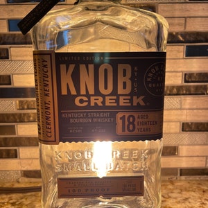 Może przedstawiać: Przezroczysta butelka szklana z brązową etykietą, na której widnieje napis "Knob Creek Kentucky Straight Bourbon Whiskey" oraz "Dojrzewany przez osiemnaście lat". Butelka ma 100 proof i jest limitowaną edycją. Butelka stoi na drewnianej podstawie.