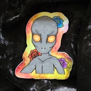 May include: A sticker featuring a gray alien with large orange eyes and a small mouth. The alien is surrounded by colorful flowers, including red, blue, and purple roses. The background has a yellow and orange gradient.