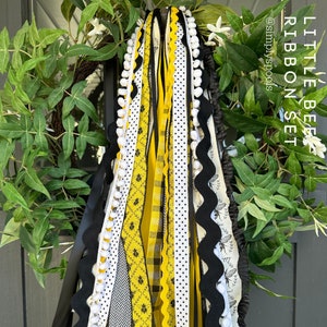 May include: A set of yellow and black ribbon streamers with white polka dots, bee patterns, and pom-poms. The ribbons are tied together and hang from a wreath. The text "LITTLE BEE RIBBON SET @simplyspools" is visible in the image.