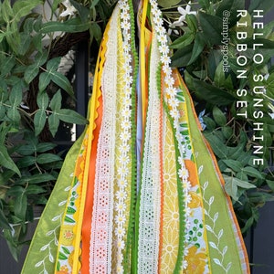 May include: A set of colorful ribbons with a daisy and orange motif. The ribbons are tied together and have a green pom-pom accent. The ribbons are labeled "Hello Sunshine Ribbon Set" and "@simplyspools".