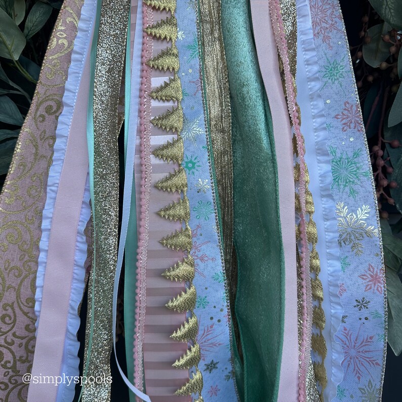 Op de afbeelding: Een kerstlintengirlande met een verscheidenheid aan linten in roze, groen, goud en wit met sneeuwvlok- en boompatronen. De guirlande is gemaakt van verschillende texturen, waaronder glitter, fluweel en satijn.