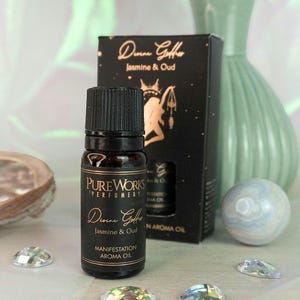 Könnte beinhalten: Eine dunkle Glasflasche "Divine Gofre Jasmine & Oud Manifestation Aroma Oil" mit schwarzem Verschluss. Eine schwarze Schachtel mit goldfarbenen Buchstaben und eine dekorative grüne Vase befinden sich hinter der Flasche. Dekorative Edelsteine sind um die Flasche herum verstreut.