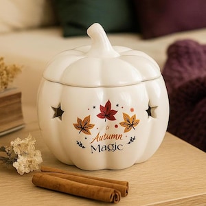May include: A white ceramic pumpkin-shaped container with a lid, featuring star-shaped cutouts and the text "Autumn Magic" with leaf illustrations. Cinnamon sticks rest on a wooden surface next to the container.