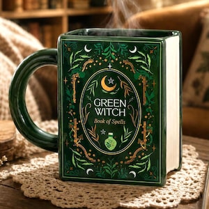 Könnte beinhalten: Ein rechteckiger, dunkelgrüner Keramikbecher in Buchform mit einem geschwungenen Henkel. Die Vorderseite zeigt goldene und weiße Illustrationen von Blättern, Sternen und einer Mondsichel sowie den Text "GREEN WITCH Book of Spells" und eine kleine Flasche.