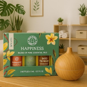 Könnte beinhalten: Eine grüne Box mit ätherischen Ölen mit der Aufschrift "Happiness" mit drei kleinen Flaschen: "Sunshine", "Meditate" und "Into The Woods". Im Vordergrund befindet sich ein ätherischer Öl-Diffusor aus Holz. Die Box ist mit gelben Blumen und Blättern verziert.