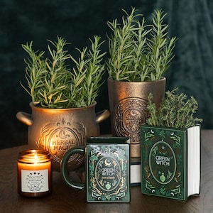 Herbal Magic Kräutertopf im Kessel-Design aus Terrakotta mit Rosmarin, dekoriert mit Kerze und Green Witch Tasse auf Holztisch