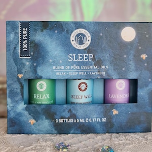 Könnte beinhalten: Eine blaue Box mit einem Mond- und Sternenmuster enthält drei kleine Flaschen mit ätherischen Ölmischungen. Die Flaschen sind mit "Relax", "Sleep Well" und "Lavender" beschriftet. Die Box trägt außerdem die Aufschrift "100% Pure", "Song of India", "Since 1932", "Sleep", "Blend of Pure Essential Oils" und "3 Bottles x 5 ml c 0.17 fl oz".
