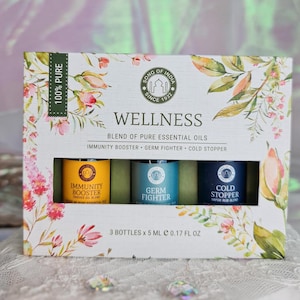 Könnte beinhalten: Ein Set mit drei 5 ml-Flaschen ätherischer Öle in einer weißen Schachtel mit Blumenmuster. Die Flaschen sind mit "Immunity Booster", "Germ Fighter" und "Cold Stopper" beschriftet. Die Schachtel trägt außerdem die Aufschrift "Wellness Blend of Pure Essential Oils" und "100% Pure".