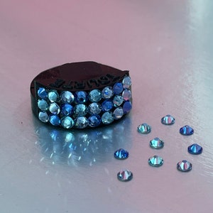 May include: A black ring with a blue, teal and purple ombre crystal design. The ring is surrounded by loose crystals of the same colours.