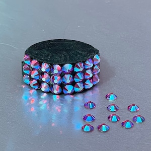 May include: A black round base with a single row of iridescent purple rhinestones around the edge. There are several loose iridescent purple rhinestones scattered on a silver surface.