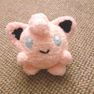 May include: Pink plush toy with black ears and blue eyes. The toy is shaped like a round, fluffy creature with a small smile.