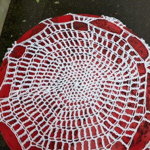 Può includere: Tovaglietta all'uncinetto bianca su un piano da tavolo rosso. La tovaglietta è circolare e ha un disegno a ragnatela.