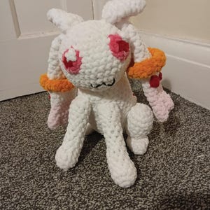 May include: A handmade white plush bunny with pink and orange accents. The bunny has large pink eyes, orange armbands, and pink paws. The plush toy is made of soft, textured yarn and is sitting on a patterned carpet.