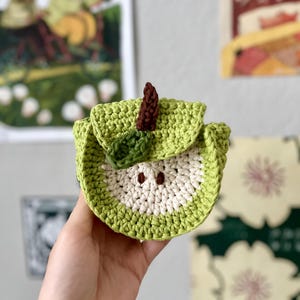 Puede incluir: Una bolsa con forma de manzana tejida a ganchillo en tonos verdes, blancos y marrones. La bolsa se sostiene en una mano y tiene un cierre de solapa. El diseño imita la apariencia de una manzana cortada, con un tallo marrón.