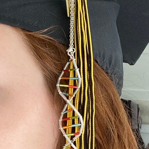 May include: A graduation cap tassel with a DNA strand charm. The tassel is black, yellow, and gold. The DNA charm is silver with colorful accents. The tassel is attached to a black graduation cap.
