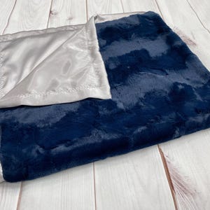 May include: A navy blue and silver baby blanket. The blanket has a plush, textured navy blue side and a smooth, satin silver side. The edges are finished with a silver satin trim.