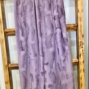 May include: A soft, plush throw blanket in a light purple color with a textured, swirl pattern. The blanket is draped over a wooden ladder.