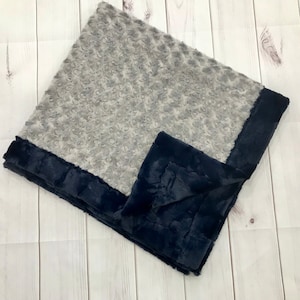 May include: A soft, gray and navy blue baby blanket with a textured, rosette-like pattern. The blanket is folded in half and has a navy blue border.