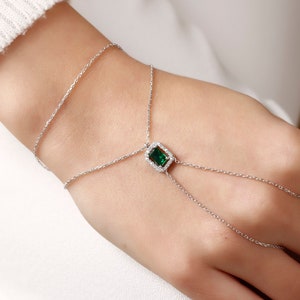 Peut inclure: Un bracelet en chaîne argent avec une pierre précieuse verte sertie dans un cadre carré. Le bracelet a une boucle de doigt qui s'étend du poignet.