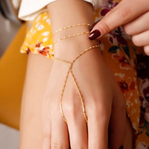May include: A gold chain bracelet with a delicate design. The bracelet is connected to a ring that can be worn on the finger. The bracelet is worn on a person's wrist.