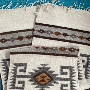 May include: Off-white woven textile with geometric patterns in shades of gray, brown, and tan. The design includes diamond and zigzag motifs, with fringed edges. The textiles are likely used as decorative elements.