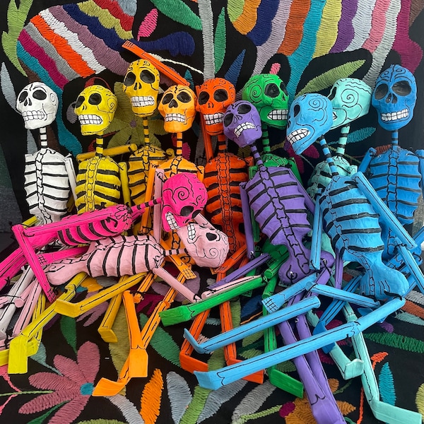 May include: A collection of articulated skeleton figurines in various vibrant colors including white, yellow, orange, pink, purple, green, and blue. Each skeleton features hand-painted black details on the skull and ribcage, with movable limbs.