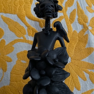 May include: A black Day of the Dead figurine with a skull head, floral headdress, and a bird on its shoulder. The figurine is adorned with a floral skirt. The background is a white fabric with yellow floral embroidery.