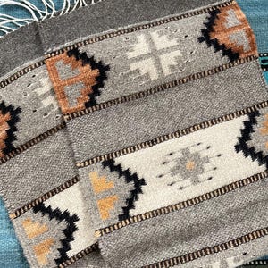 May include: A folded gray textile with a geometric pattern in black, tan, and white. The textile has a fringed edge and is layered over a turquoise textile with a patterned border. The design features repeating shapes.