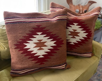 May include: Two square throw pillows with a geometric design. The pillows have a reddish-brown background with a central diamond pattern in white, red, and brown. The top border features stripes of white, red, and brown.