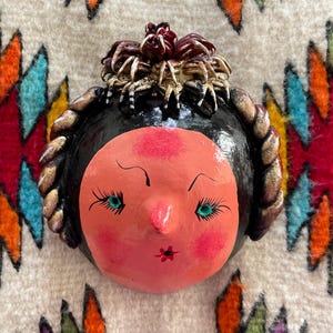 May include: A hand-painted ceramic face with a pink complexion, black hair, and teal eyes. The face has a small red flower on the lips and is adorned with a decorative headpiece. The background is a colorful patterned textile.