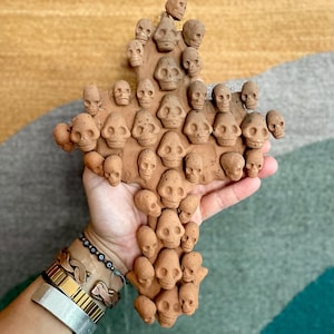 May include: A terracotta cross made of many small, detailed skull shapes. The cross is held in a hand with several bracelets on the wrist.