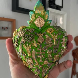 May include: A green heart-shaped decorative object adorned with various gold and silver-toned religious charms. The heart features a gold medallion and a leaf-like design at the top. The object is held in a hand.