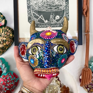 Może przedstawiać: Kolorowa, ręcznie malowana maska o diabelskim wzorze. Posiada złote rogi, duże oczy i wystający różowy język. Podstawą jest niebieski kolor, ozdobiony kwiatowymi wzorami. Maska trzymana jest na tle ściany z oprawionymi dziełami sztuki.