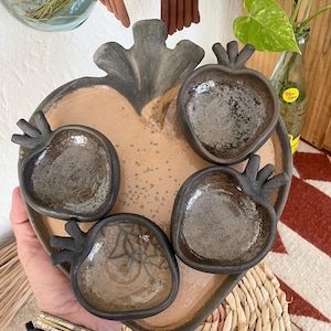 Op de afbeelding: Een handgemaakt keramisch hartvormig serveerblad met vier kleine, appelvormige kommen. De bakplaat is bruin met een donkergrijze rand en de kommen zijn donkergrijs.