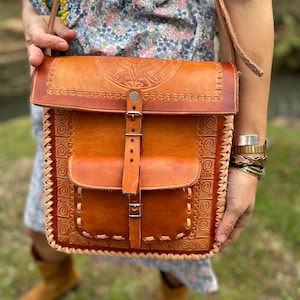 May include: A brown leather crossbody bag with intricate embossed designs and a flap closure with a buckle. The bag has a smaller pocket on the front and a braided leather trim around the edges.