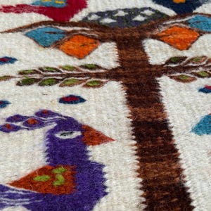 May include: Close-up of a woven textile featuring a tree design with colorful birds. The birds are red, blue, and purple, with orange and green accents. The tree trunk is brown, and the background is off-white.