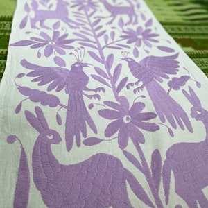 May include: A white table runner with a purple embroidered design featuring birds, flowers, and deer. The design is traditional Mexican folk art.
