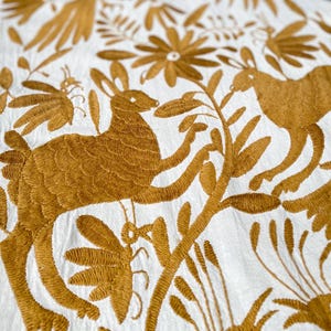 Puede incluir: Primer plano de tela blanca con intrincado bordado dorado. El diseño presenta conejos, pájaros, flores y follaje. La costura detallada crea un patrón texturizado, ideal para textiles o decoración del hogar.