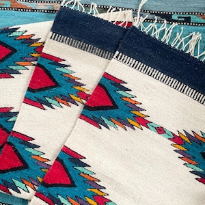 Peut inclure: Textile rectangulaire tissé blanc avec un motif géométrique rouge, turquoise, jaune et violet. Le bord supérieur est bleu marine avec une frange blanche. Le textile est posé sur un fond tissé turquoise et bleu.