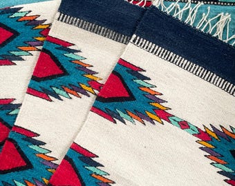 May include: White, rectangular woven textile with a geometric design in red, turquoise, yellow, and purple. The top edge is navy blue with a white fringe. The textile is placed on a turquoise and blue woven background.