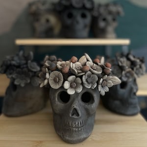 May include: Several dark gray ceramic skull figurines, each adorned with a floral crown. The skulls have open eye sockets and a smiling mouth. The flowers are in shades of cream, brown, and gray.
