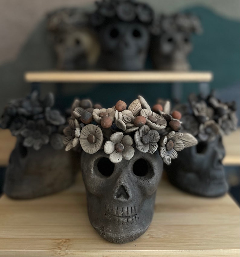 Pode incluir: V&aacute;rias figuras de caveiras de cer&acirc;mica cinza escuro, cada uma adornada com uma coroa floral. As caveiras t&ecirc;m &oacute;rbitas oculares abertas e uma boca sorridente. As flores s&atilde;o em tons de creme, castanho e cinza.