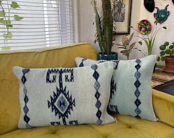 May include: Two light blue throw pillows with a black and white geometric pattern on a mustard yellow couch.
