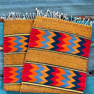 May include: Two rectangular woven placemats with a mustard yellow background and a vibrant zigzag pattern in red, orange, and blue. The placemats have a fringed edge and are displayed on a turquoise woven surface.