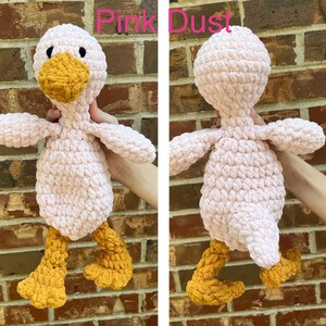 May include: Pink crocheted duck plush toy with yellow feet and beak. The duck is standing on a brick wall.